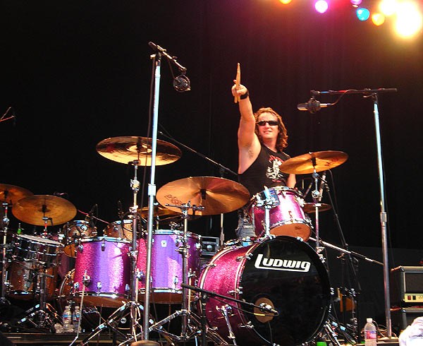 Randy Lane pointing skyward behind his Ludwig kit with Paiste cymbals on an outdoor stage
